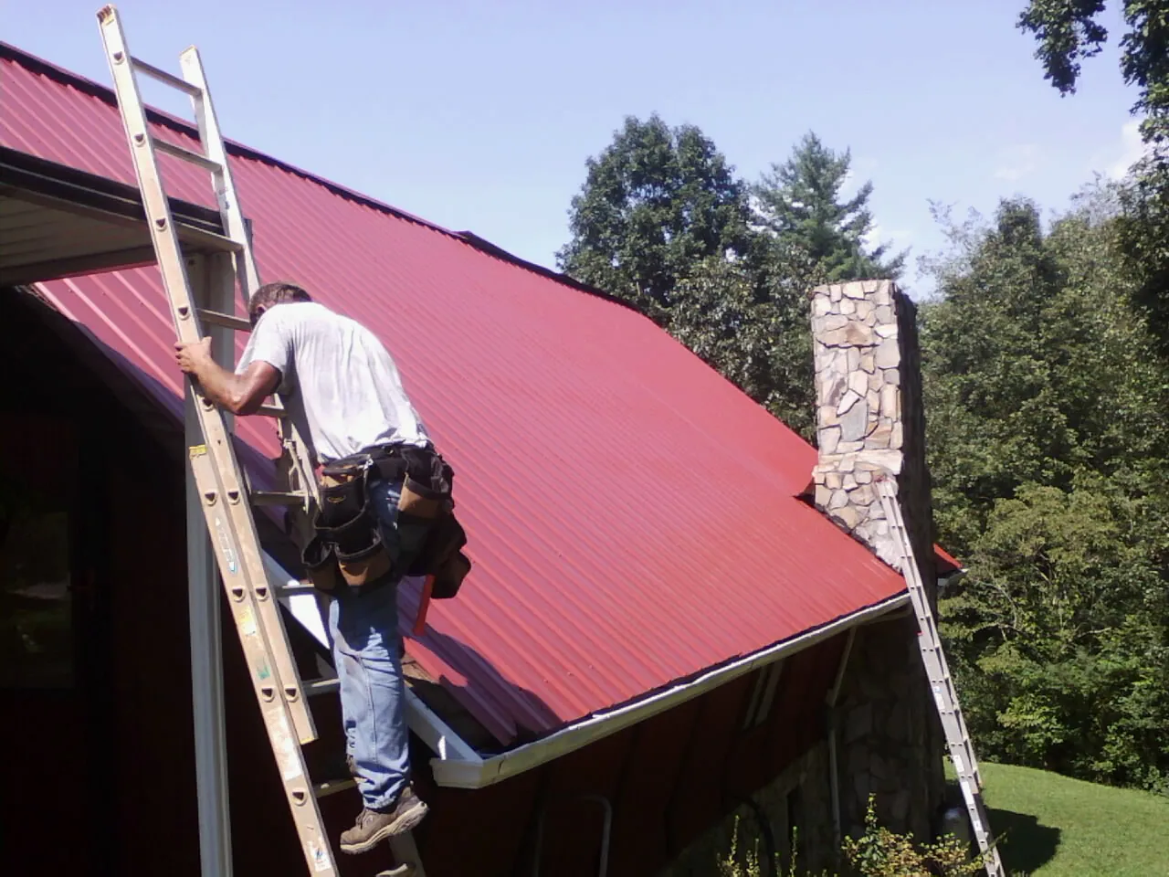 Roofer on a ladder beside a red metal roof and stone chimney — Soffit & Fascia Repair by Roofing Brothers in Dowling